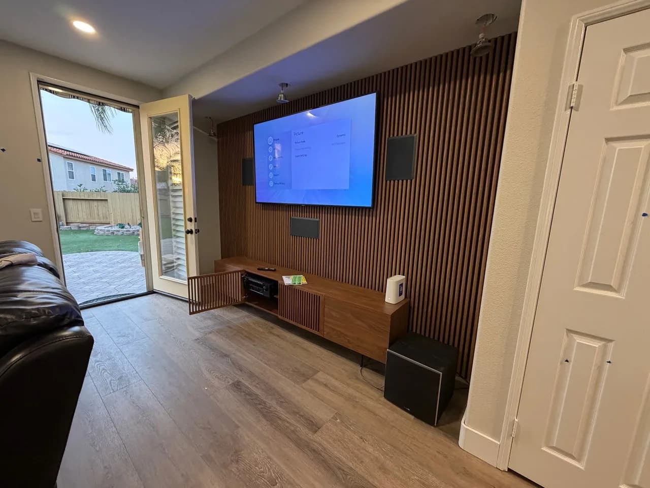 A completed wood-slat media wall with a mounted television, three wall-mounted speakers, a media console, and a floor-standing subwoofer.